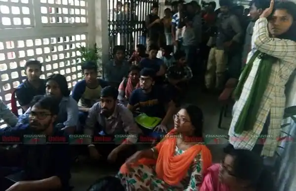 শিক্ষার্থীরা ভিসি কার্যালয়ের সামনে বিক্ষোভ করছে, অনেক ছাত্র-ছাত্রী বসে ও দাঁড়িয়ে. Channel i News - চ্যানেল আই নিউজ