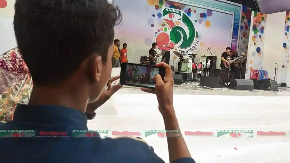 একজন ব্যক্তি স্টেজে পারফর্মারদের ছবি মোবাইলে ধারণ করছে. Channel i News - চ্যানেল আই নিউজ