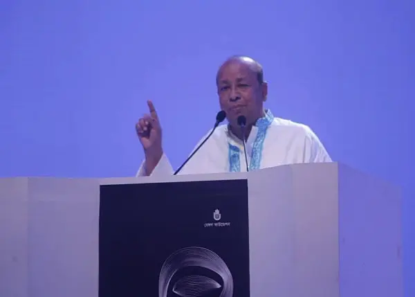 বাংলাদেশের একজন বক্তা মঞ্চে বক্তৃতা দিচ্ছেন, পেছনে নীল ব্যাকগ্রাউন্ড।. Channel i News - চ্যানেল আই নিউজ