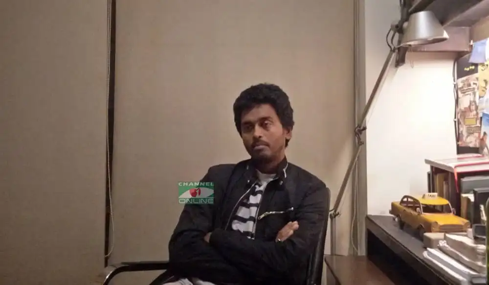 একজন যুবক বসে আছেন, পেছনে একটি ডেস্ক ও কিছু বই ও গাড়ির মডেল. Channel i News - চ্যানেল আই নিউজ