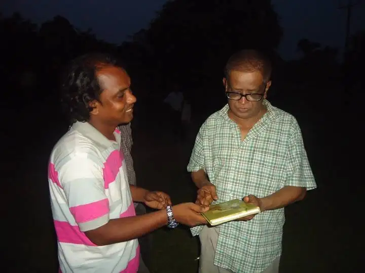 দুই ব্যক্তি বই পড়ছেন, একজনের হাতে বই, সন্ধ্যার সময়. Channel i News - চ্যানেল আই নিউজ