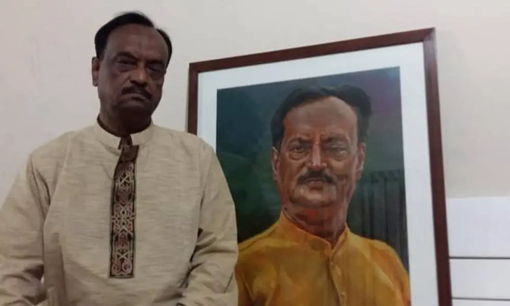 একজন ব্যক্তি ছবি পাশে দাঁড়িয়ে, পেছনে বড় পোর্ট্রেটের ছবি রয়েছে।. Channel i News - চ্যানেল আই নিউজ