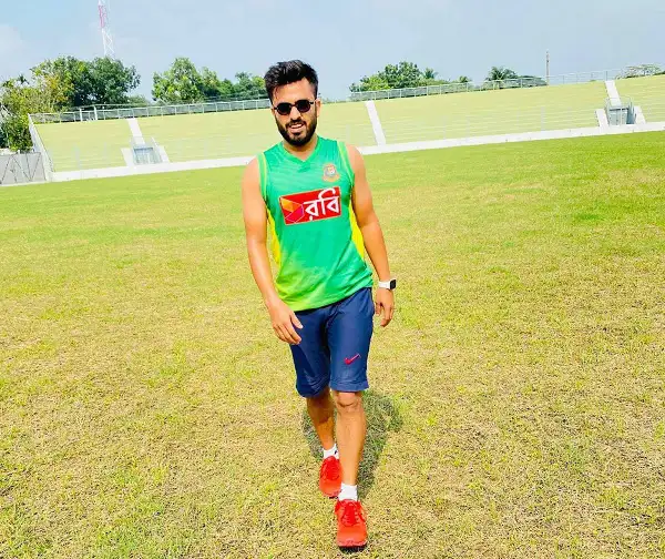 বাংলাদেশি ক্রিকেটার মাঠে হাঁটছেন, সবুজ মাঠের পেছনে দর্শক বসে. Channel i News - চ্যানেল আই নিউজ