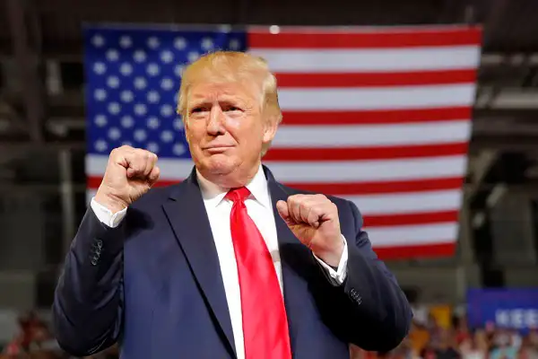 Donald Trump at a rally with American flag backdrop, making a fist gesture. Channel i News - চ্যানেল আই নিউজ