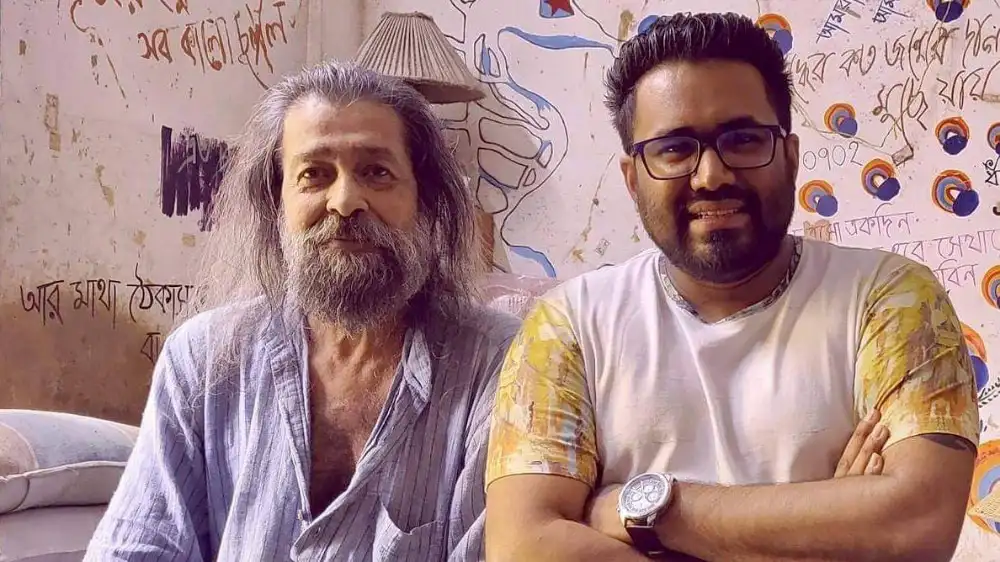 দুই ব্যক্তি হাস্যোজ্জ্বলভাবে দাঁড়িয়ে আছেন, পেছনে দেয়াল ও চিত্রাঙ্কন দেখা যাচ্ছে. Channel i News - চ্যানেল আই নিউজ