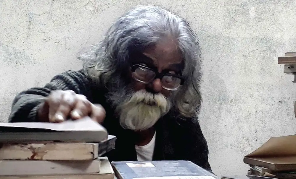 প্রবীণ ব্যক্তি বই পড়ছেন, চোখে চশমা, পেছনে সাদা দেয়াল. Channel i News - চ্যানেল আই নিউজ