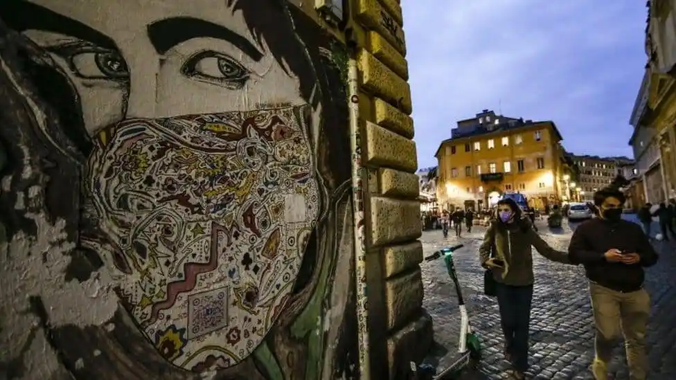 Street scene in Italy with people wearing masks and a mural of a face with a mask. Channel i News - চ্যানেল আই নিউজ