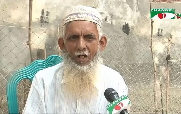 বৃদ্ধ মুক্তিযোদ্ধা কথা বলছেন, পেছনে ধাতব জাল ও দেয়াল দেখা যাচ্ছে।. Channel i News - চ্যানেল আই নিউজ