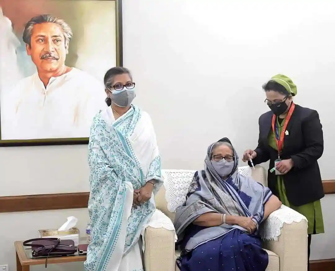 প্রধানমন্ত্রী ভ্যাকসিন গ্রহণের সময় একজন নারীর পাশে অন্য নারী ও একজন নার্স দাঁড়িয়ে. Channel i News - চ্যানেল আই নিউজ