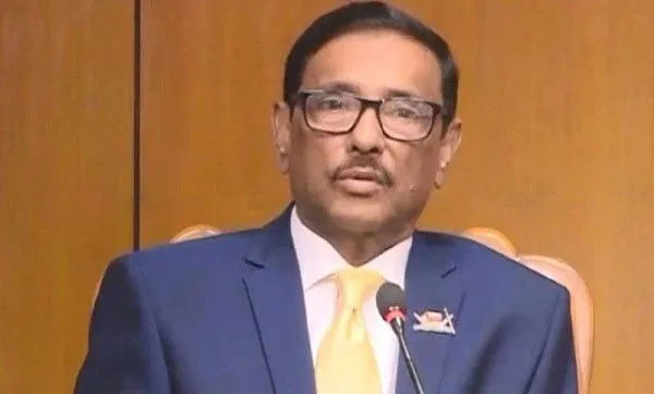 ওবায়দুল কাদের speaking at a press conference in a blue suit and yellow tie. Channel i News - চ্যানেল আই নিউজ