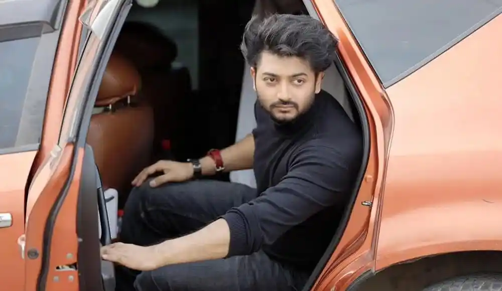 বাপ্পী গাড়ির ভিতরে বসে পোজ দিচ্ছেন, কালো পোশাক পরা. Channel i News - চ্যানেল আই নিউজ