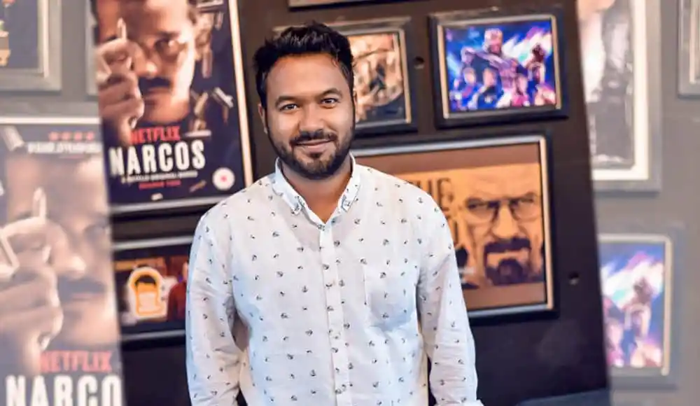 একজন হাস্যোজ্জ্বল পুরুষের ছবি, পটভূমিতে টেলিভিশন ও সিনেমার পোস্টার. Channel i News - চ্যানেল আই নিউজ