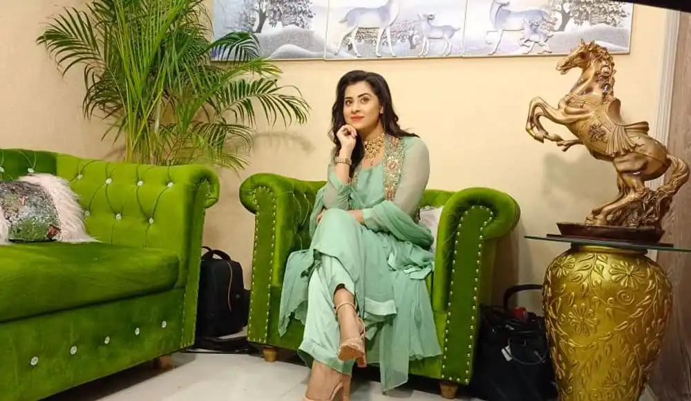 বুবলী বসে আছেন একটি সবুজ সোফির উপর, পেছনে গাছ ও সোনালী ভাস্কর্য. Channel i News - চ্যানেল আই নিউজ
