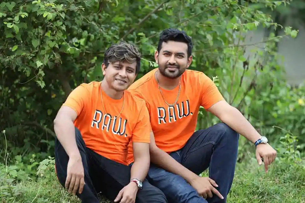দুই যুবক বসে আছেন সবুজ গাছের পাশে, হাসিমুখে. Channel i News - চ্যানেল আই নিউজ
