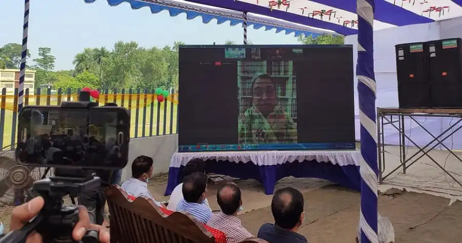 Bangladeshi officials watch a virtual event on a large screen outdoors. Channel i News - চ্যানেল আই নিউজ