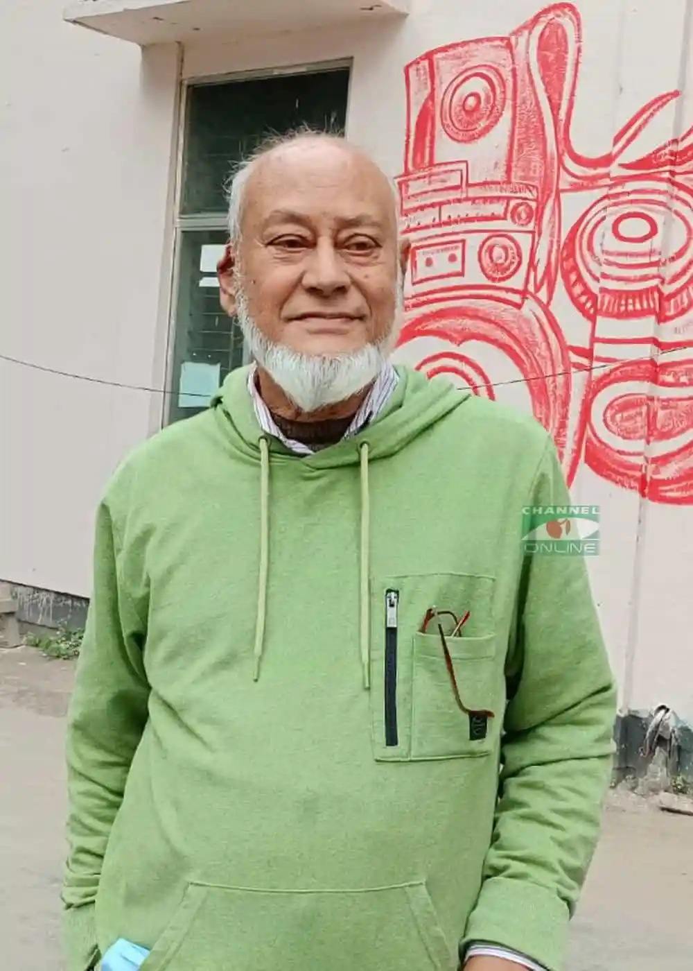বয়স্ক ব্যক্তি হালকা সবুজ হুডি পরা, হাসিমুখে দাঁড়িয়ে. Channel i News - চ্যানেল আই নিউজ