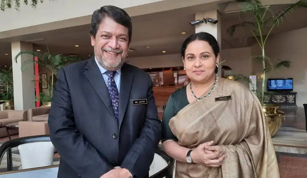 দুটি মানুষ হাসিমুখে একসঙ্গে দাঁড়িয়ে আছেন, অফিস বা হোটেল লাউঞ্জে।. Channel i News - চ্যানেল আই নিউজ