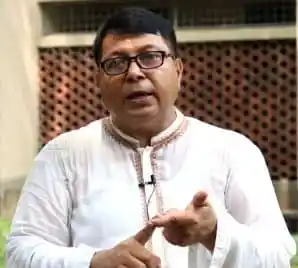 বাংলাদেশের একজন ব্যক্তি কথা বলছেন, পর্দার পেছনে গাছের পাতা ও কাঠের ফাঁদ।. Channel i News - চ্যানেল আই নিউজ