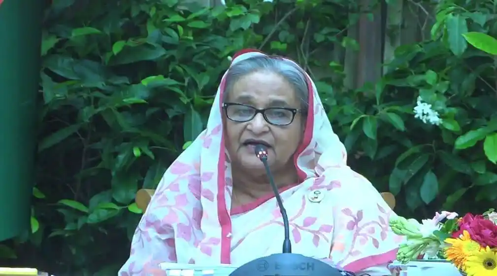 প্রধানমন্ত্রী speaking at a public event with greenery in the background. Channel i News - চ্যানেল আই নিউজ