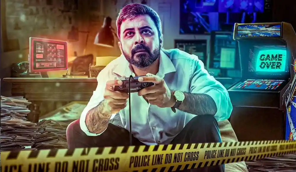 একজন পুরুষ গেমিং কনসোল হাতে নিয়ে খেলছেন, পেছনে কম্পিউটার ও মনিটর রয়েছে. Channel i News - চ্যানেল আই নিউজ
