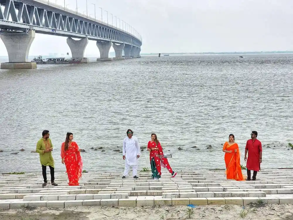 পদ্মা সেতুর পাশে নদীর পাড়ে ছয়জন মানুষ হাঁটছেন. Channel i News - চ্যানেল আই নিউজ
