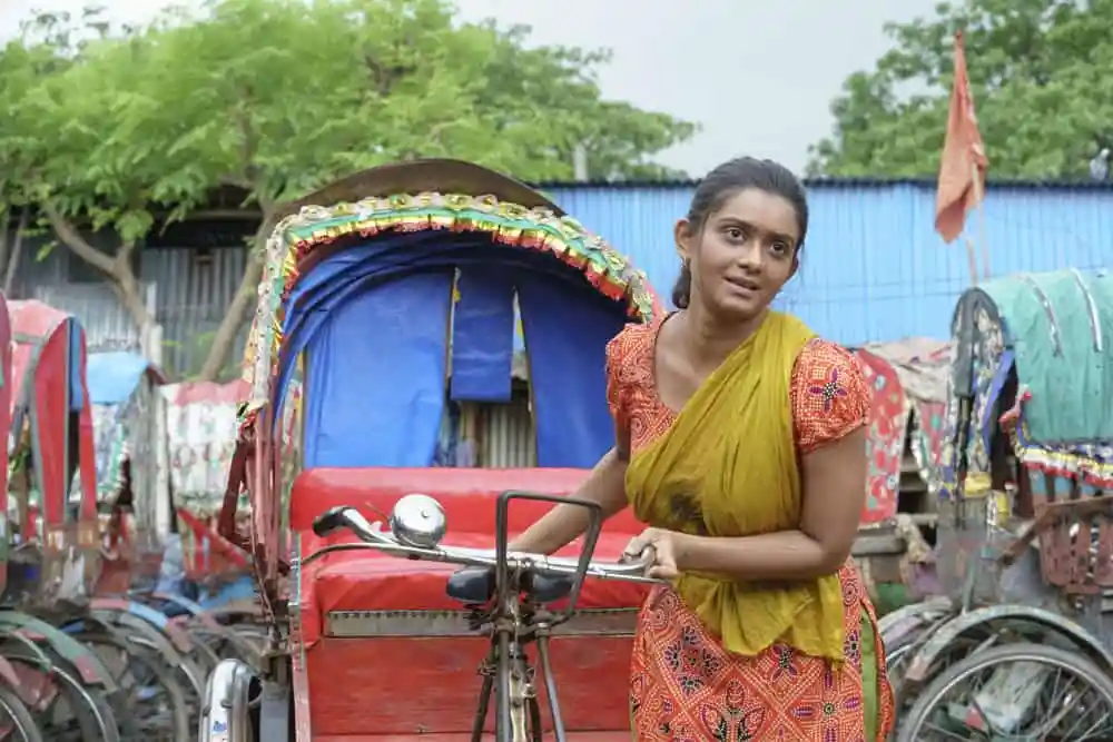 একজন নারী রিকশা চালাচ্ছেন, পেছনে রিকশা ও ট্রাক দেখা যাচ্ছে. Channel i News - চ্যানেল আই নিউজ