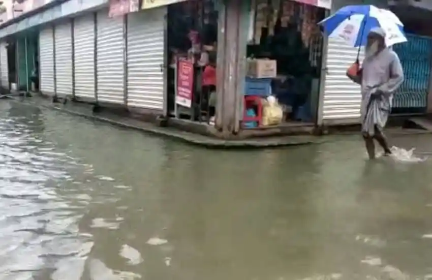 বন্যার পানিতে ডুবে গেছে দোকানপাট ও রাস্তাঘাট, একজন ব্যক্তি ছাতা হাতে হাঁটছেন।. Channel i News - চ্যানেল আই নিউজ