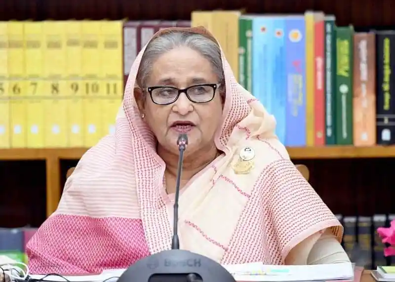 প্রধানমন্ত্রী ভাষণ দিচ্ছেন, পেছনে বইয়ের শেলফ দেখা যাচ্ছে. Channel i News - চ্যানেল আই নিউজ