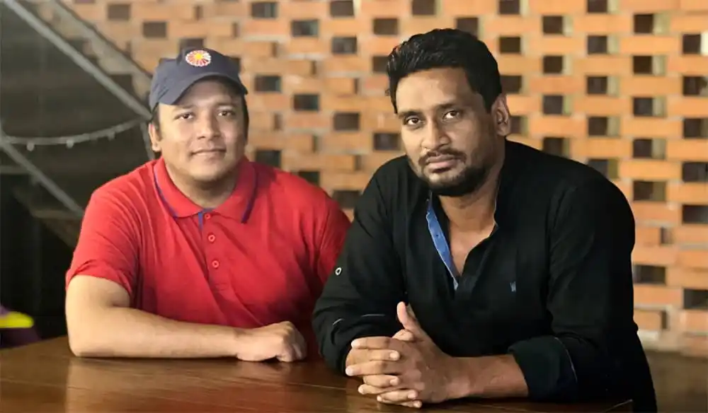 দুই ব্যক্তি বসে আছেন, একজন লাল পোশাক পরা, অন্যজন কালো পোশাক পরা. Channel i News - চ্যানেল আই নিউজ