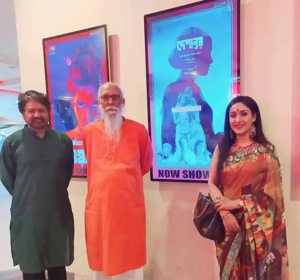 তিনজন ব্যক্তি সিনেমা পোস্টার সামনে দাঁড়িয়ে, একটি মহিলা সেজে আছেন. Channel i News - চ্যানেল আই নিউজ