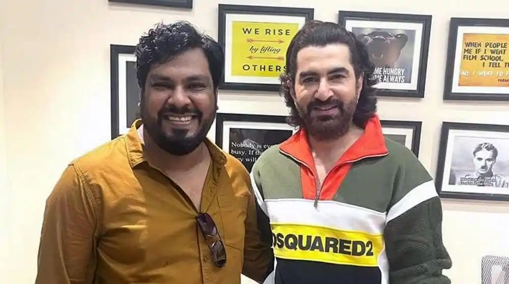 দুই ব্যক্তি হাসিমুখে দাঁড়িয়ে, পটভূমিতে বিভিন্ন পোস্টার ও ছবি. Channel i News - চ্যানেল আই নিউজ