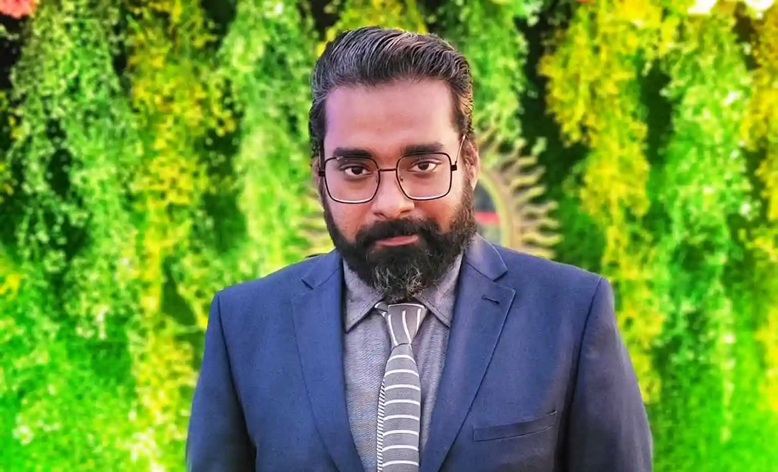 প্রতিষ্ঠানিক পোশাক পরিহিত একজন পুরুষের ছবি, পেছনে সবুজ গাছের পাতা. Channel i News - চ্যানেল আই নিউজ