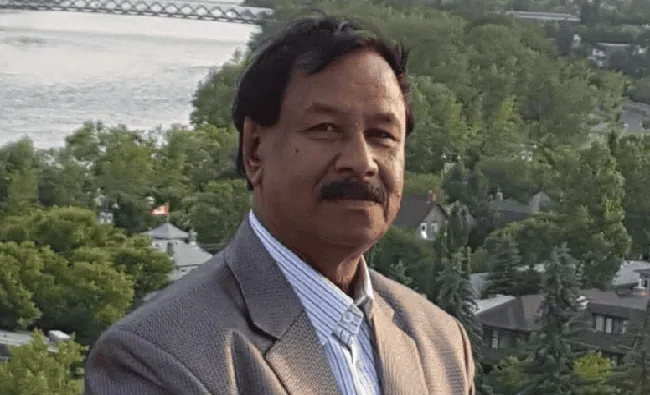 A man in a suit standing outdoors with a river and greenery in the background. Channel i News - চ্যানেল আই নিউজ