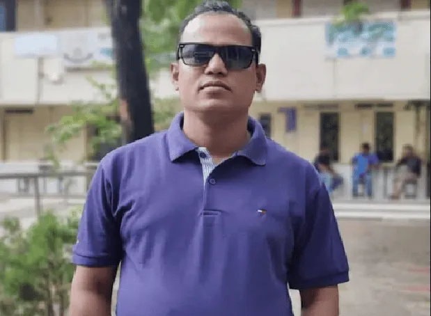 একজন ব্যক্তি কালো চশমা পরা, নীল পোশাক পরিহিত, বাইরের পরিবেশে দাঁড়িয়ে. Channel i News - চ্যানেল আই নিউজ