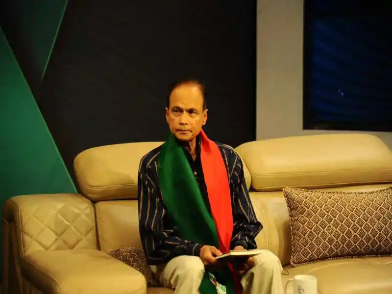 ড. মশিউর রহমান বসে থাকছেন, হাতে বই ও মোবাইল, পেছনে সোফা ও প্যানেল. Channel i News - চ্যানেল আই নিউজ
