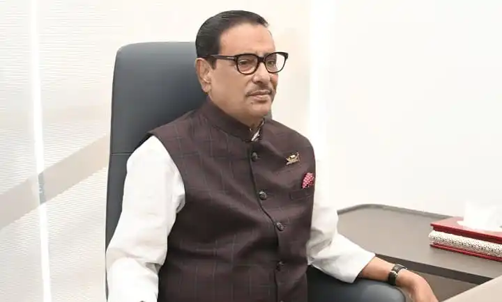 ওবায়দুল কাদের বসে আছেন, পেছনে সাদা দেয়াল ও টেবিল. Channel i News - চ্যানেল আই নিউজ
