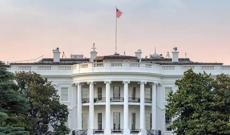 The White House with American flag flying, surrounded by trees. Channel i News - চ্যানেল আই নিউজ