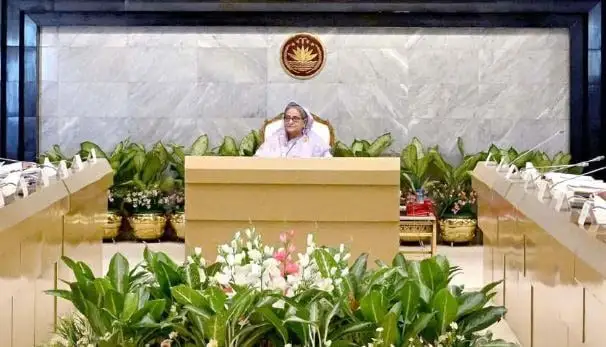 প্রধানমন্ত্রী seated at a conference table with plants and a national emblem behind. Channel i News - চ্যানেল আই নিউজ