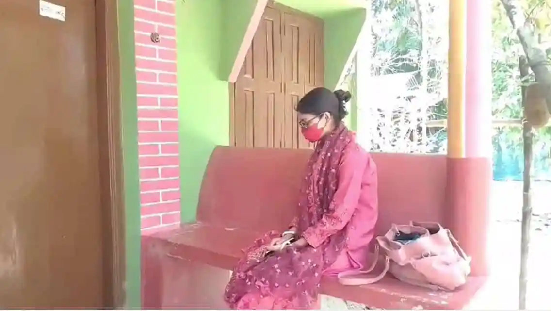নারী বসে আছেন, মুখে মাস্ক, পাশে ব্যাগ, বাড়ির বারান্দায়. Channel i News - চ্যানেল আই নিউজ