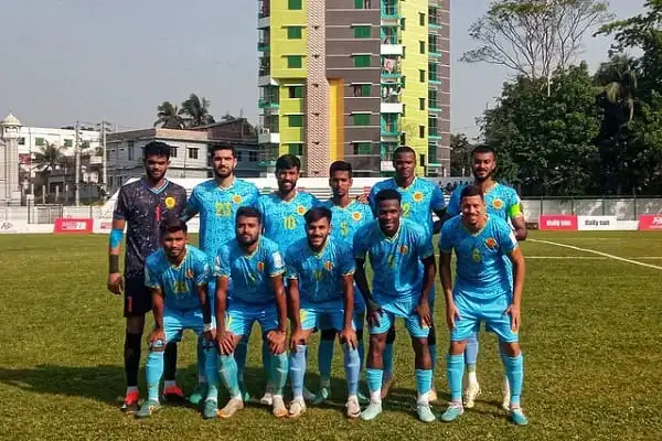 বাংলাদেশ ফুটবল দলের খেলোয়াড়রা মাঠে প্রস্তুত, পেছনে ভবন ও গাছ দেখা যাচ্ছে. Channel i News - চ্যানেল আই নিউজ