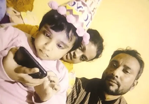 Child celebrating birthday with family, wearing party hat and looking at phone. Channel i News - চ্যানেল আই নিউজ