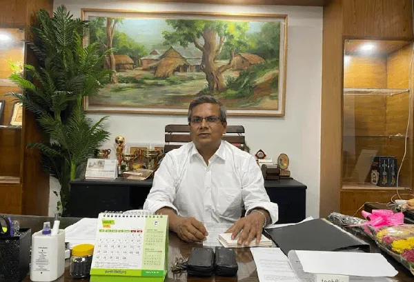 অধ্যাপক মোজাম্মেল হোসেন বসে আছেন অফিসে, পেছনে গাছ ও চিত্রকর্ম. Channel i News - চ্যানেল আই নিউজ