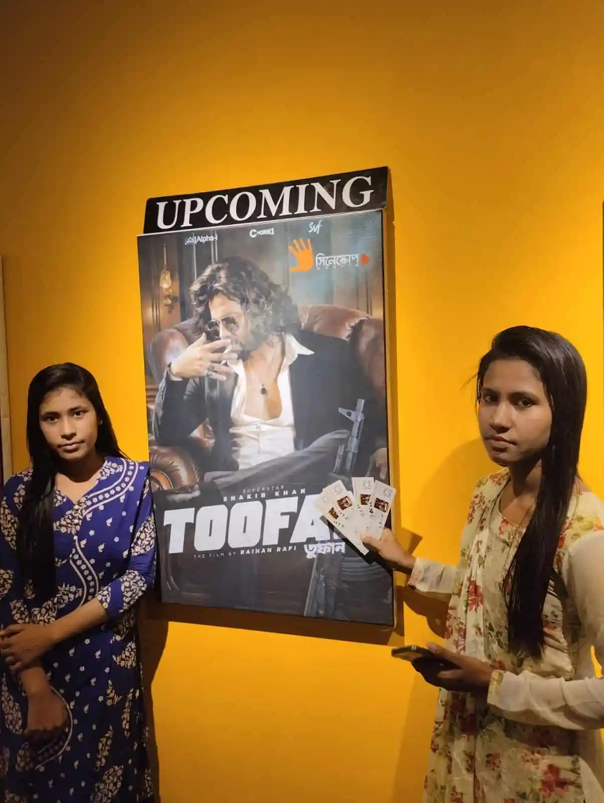 দুটি নারী দর্শক সিনেমার পোস্টার দেখছেন, একজন টিকেট ধরেছেন. Channel i News - চ্যানেল আই নিউজ