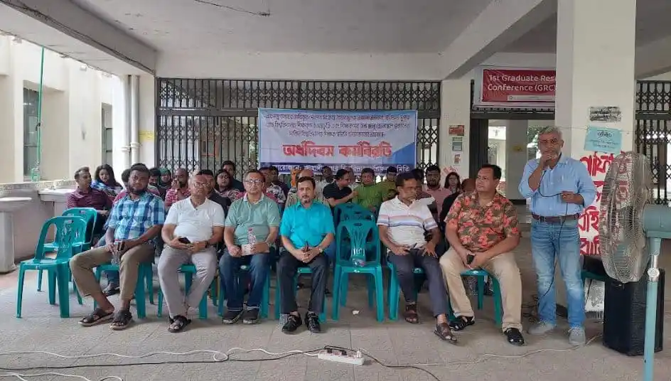 প্রতিবাদী সমাবেশে অংশগ্রহণকারীরা বসে আছেন, একটি ব্যানার পেছনে দেখা যাচ্ছে. Channel i News - চ্যানেল আই নিউজ