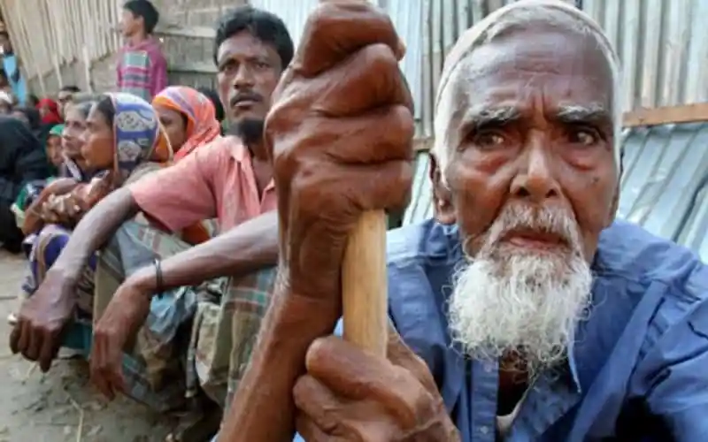 প্রবীণ ব্যক্তির হাতে লাঠি ধরে দাঁড়িয়ে আছেন, পাশে অন্যরা বসে আছেন. Channel i News - চ্যানেল আই নিউজ