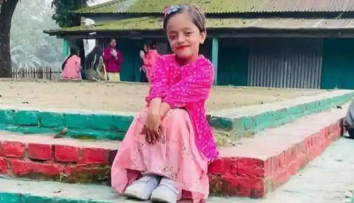 একটি ছোট মেয়ে হাসিমুখে বসে আছে, পেছনে কিছু মানুষ এবং বাড়ি দেখা যাচ্ছে. Channel i News - চ্যানেল আই নিউজ