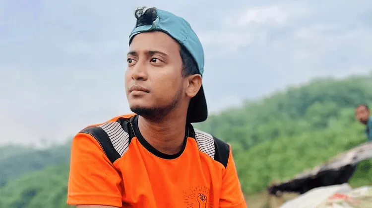 Young man in orange T-shirt outdoors with green hills in background. Channel i News - চ্যানেল আই নিউজ