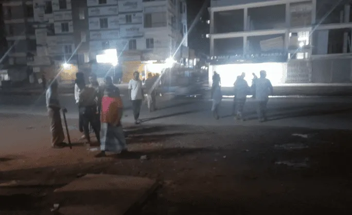 রাতের সময় ঢাকায় কিছু মানুষ রাস্তার পাশে দাঁড়িয়ে আছেন. Channel i News - চ্যানেল আই নিউজ