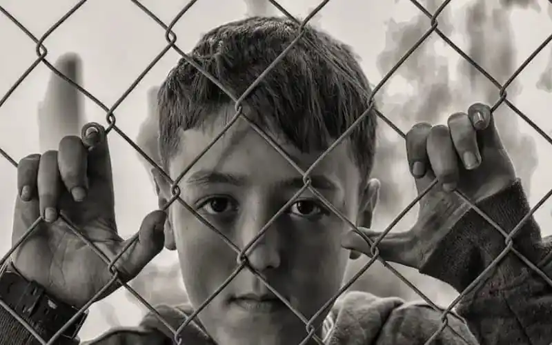 Child looking through a fence, expressing longing or sadness. Channel i News - চ্যানেল আই নিউজ