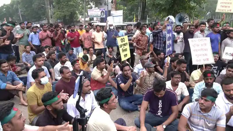 জনতা বিক্ষোভে রাস্তায় বসে, বিভিন্ন ব্যক্তি হাতে প্ল্যাকার্ড ও মাইক নিয়ে প্রতিবাদ করছে. Channel i News - চ্যানেল আই নিউজ